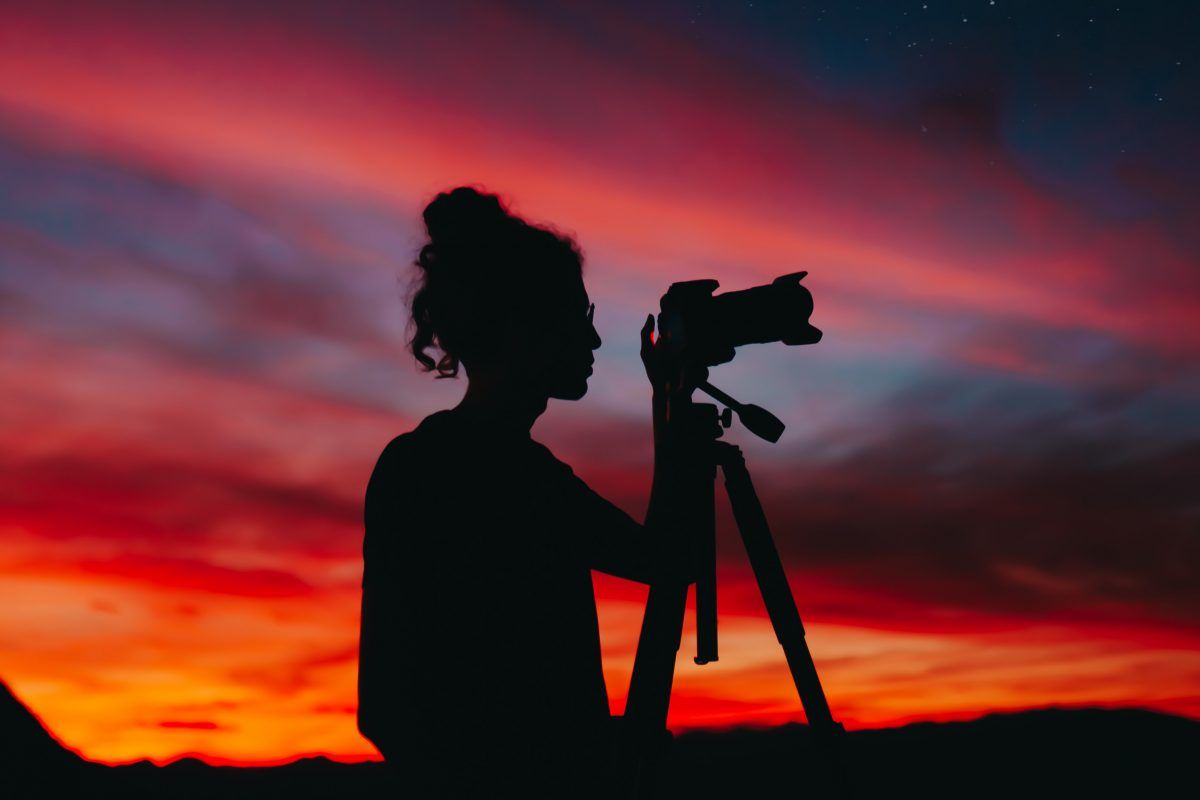 Une silhouette d'une femme tenant un appareil photo sur un trépied au coucher du soleil.