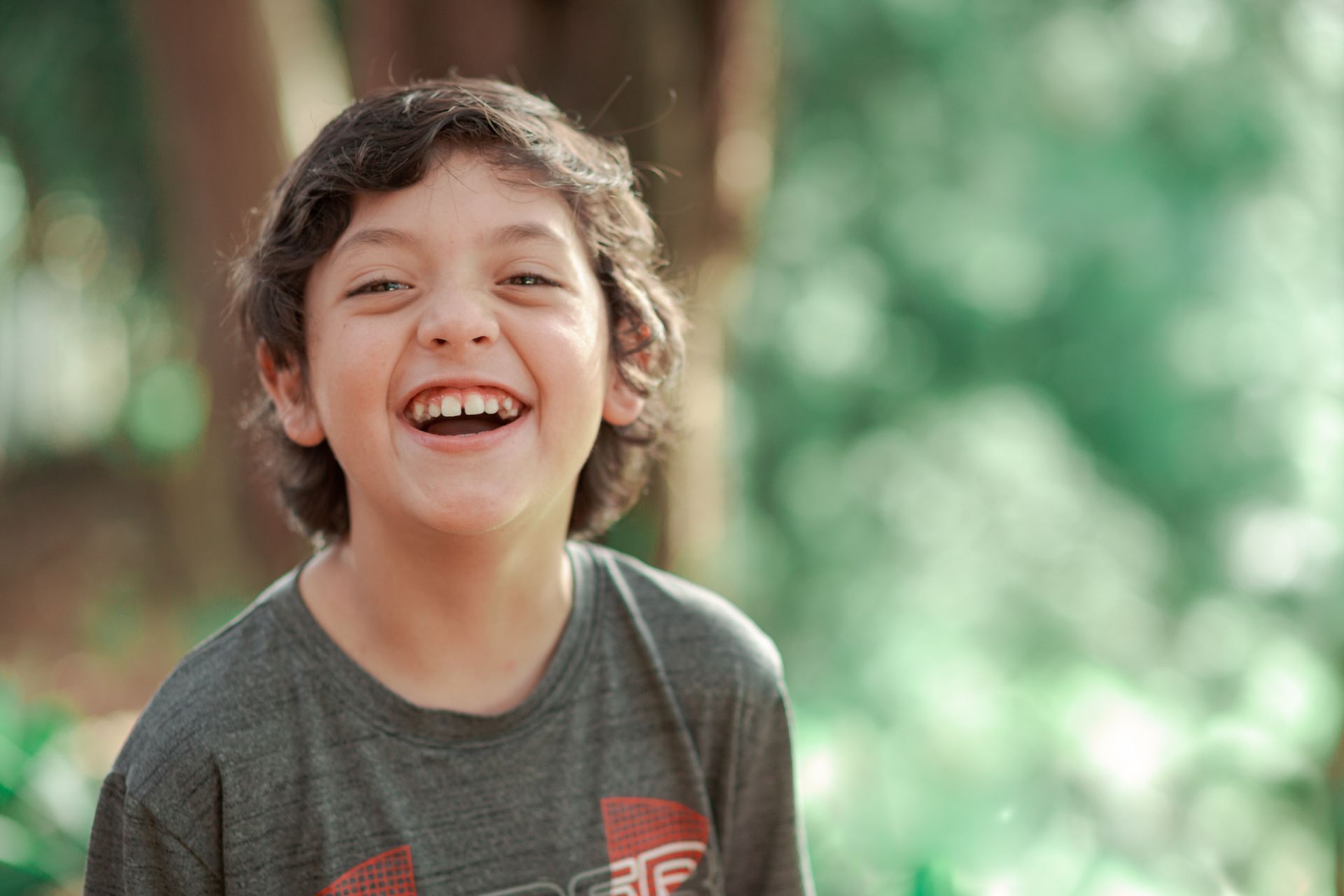 Child with long brown hair and a grey shirt, standing outdoors and smiling
