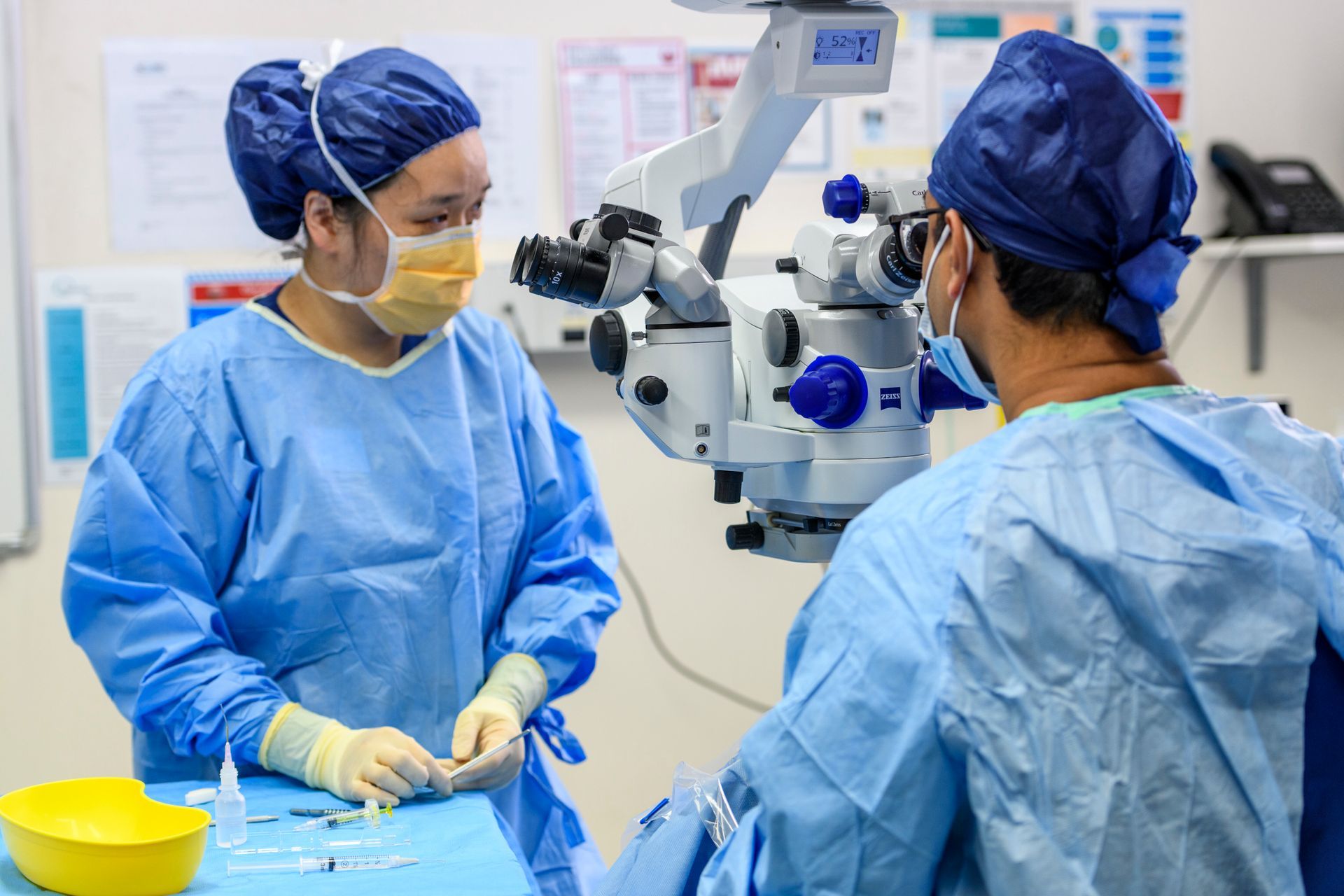Doctor in glasses and white coat, leaning on eye exam equipment, smiling in an office.