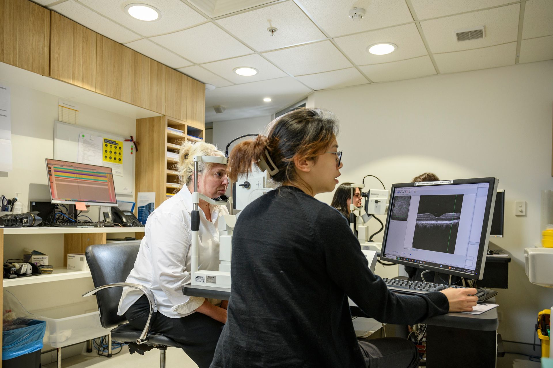 Woman in purple shirt at a reception desk; staffer in black jacket holds papers and gestures. Sydney Retina Clinic sign.