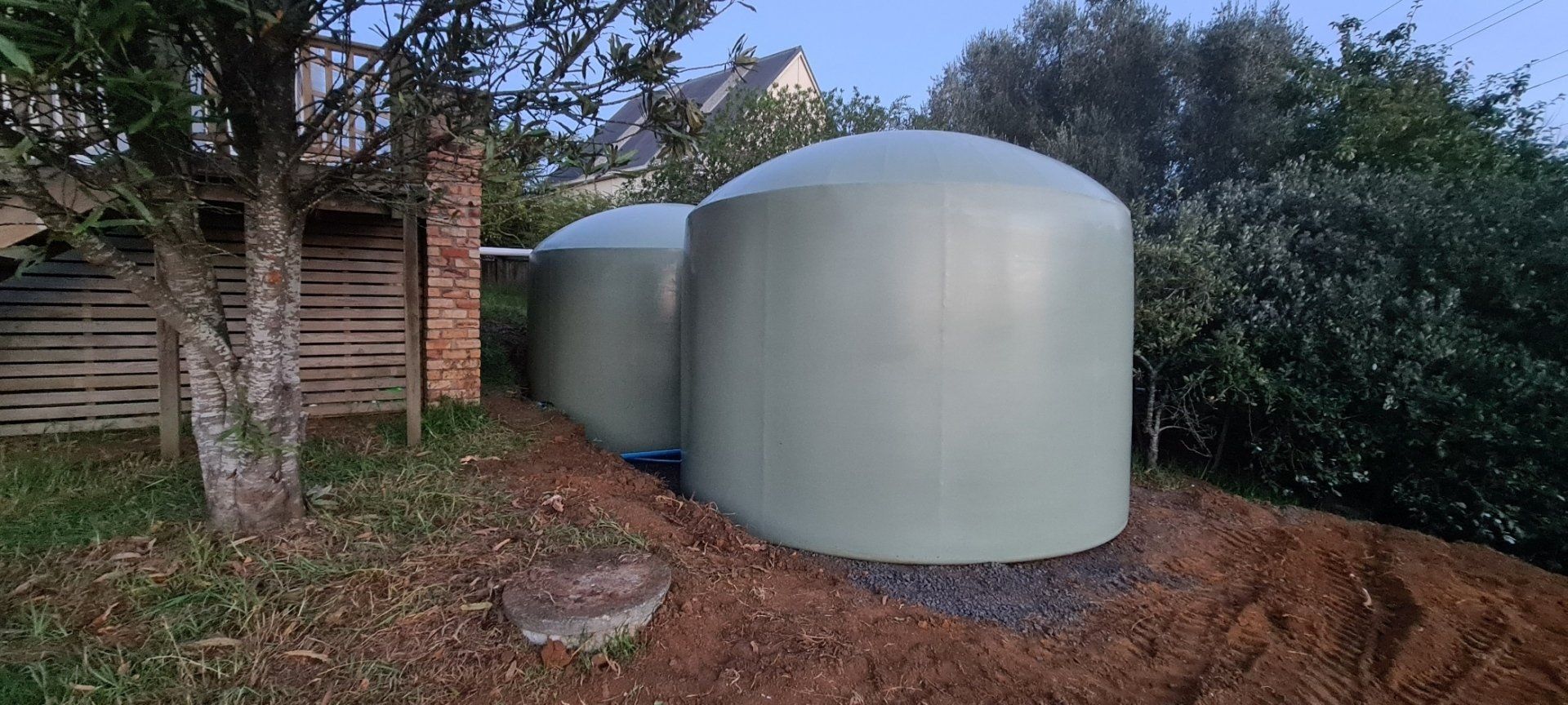 Two large, pale green tanks with rounded tops sit outside, near a fence and trees.