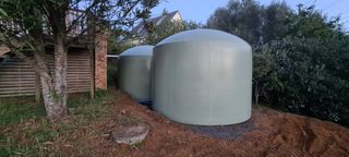 Two large, pale green tanks with rounded tops sit outside, near a fence and trees.