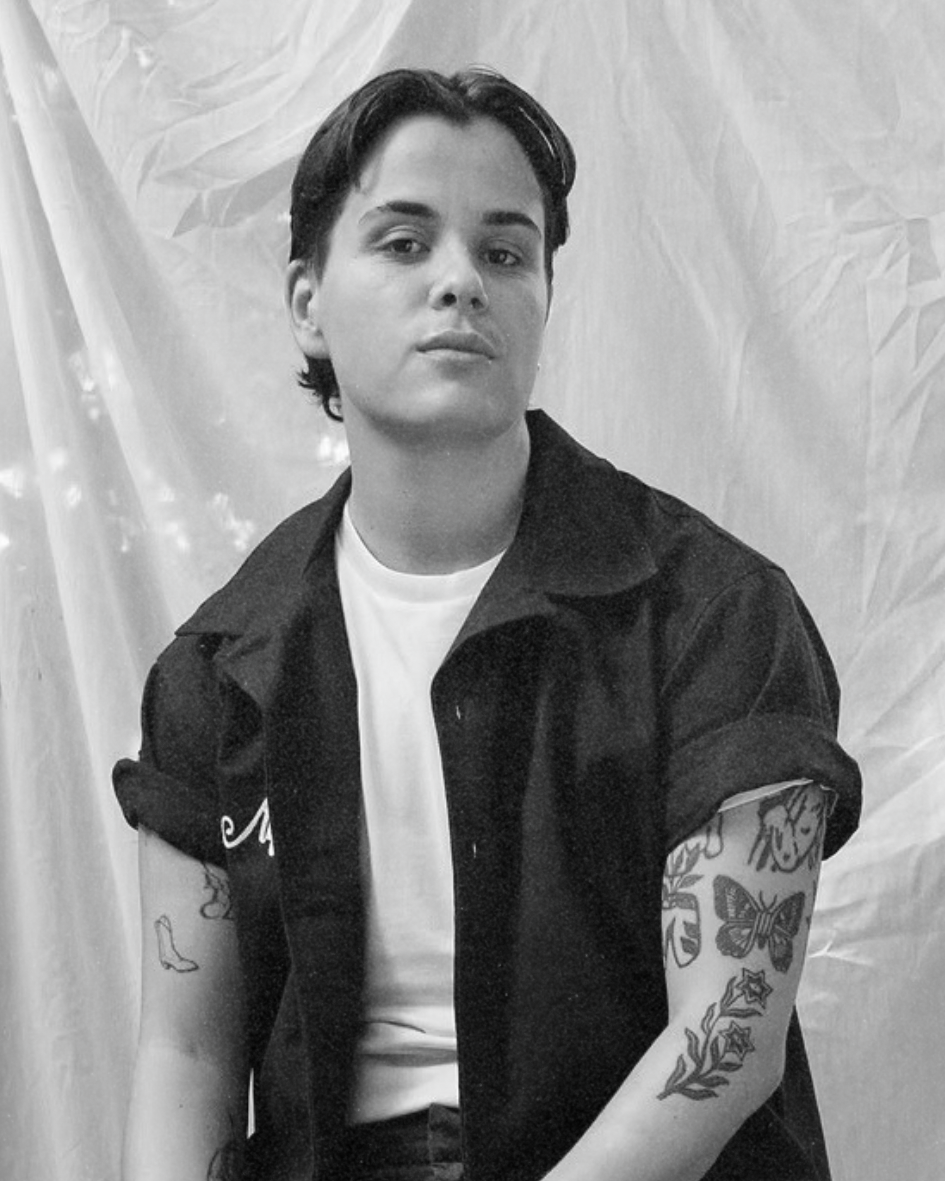 Person with short dark hair in a black shirt over a white tee, posing against a draped fabric backdrop.