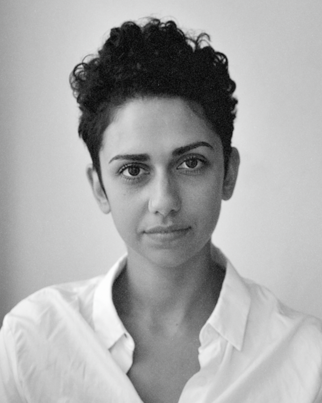 Black-and-white portrait of a person with short curly hair wearing a white collared shirt, looking at the camera
