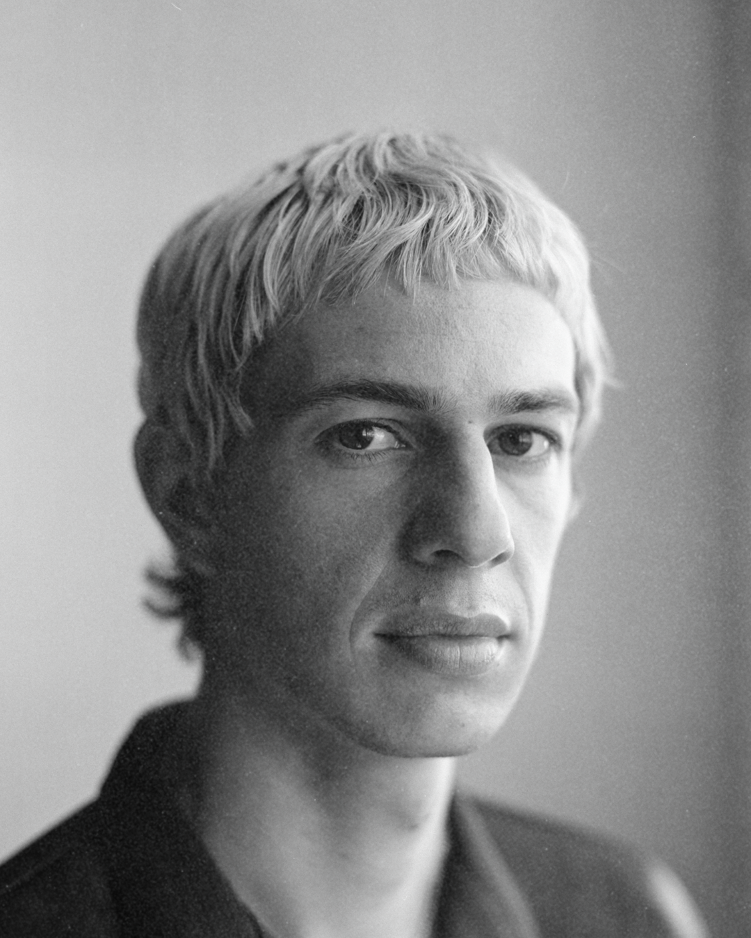 Black-and-white close-up portrait of a person with short light hair, looking at the camera.