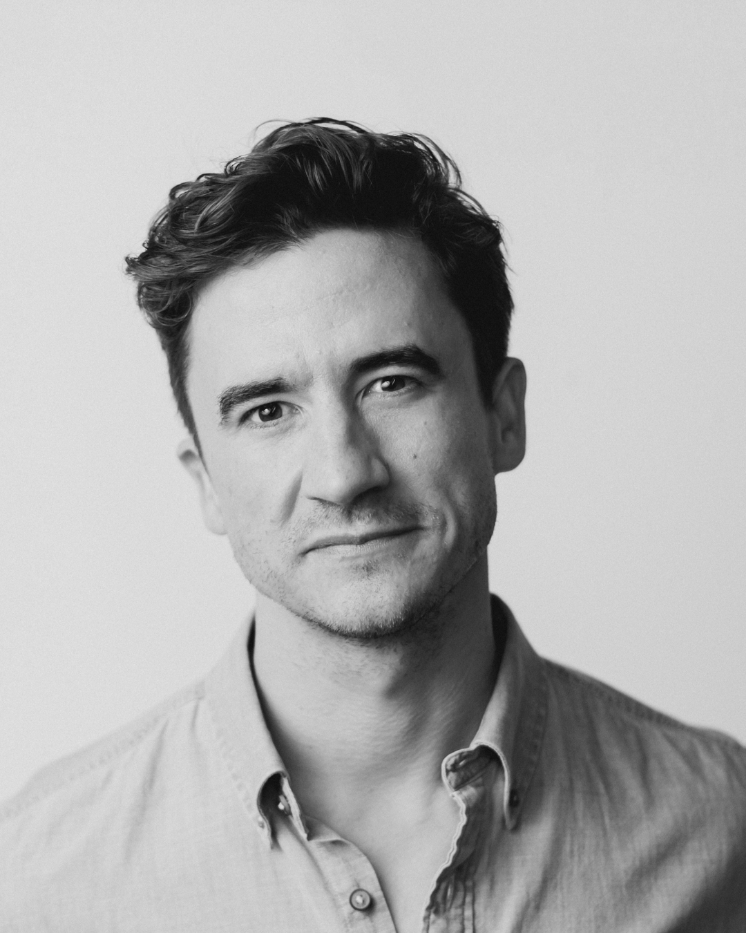Black-and-white portrait of a man with dark wavy hair, wearing an open-collar shirt, against a plain background