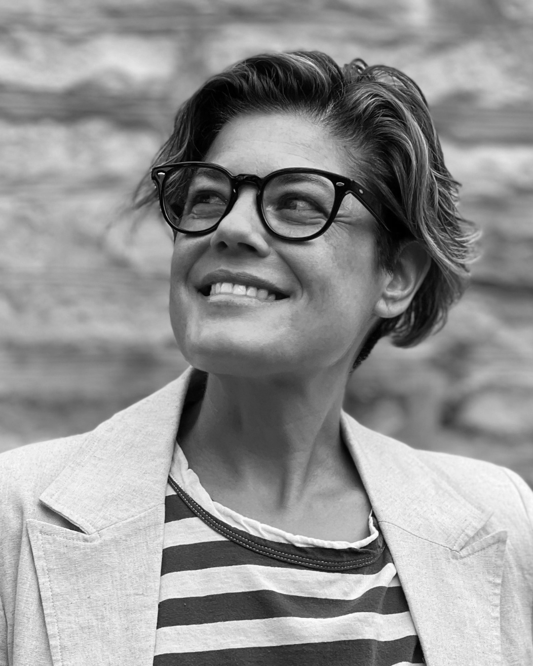 Smiling person in glasses and light blazer, looking upward against a stone wall background