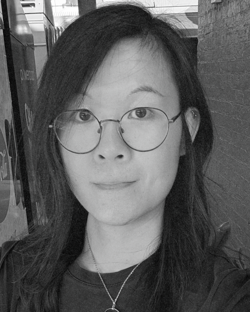 Black-and-white portrait of a person with round glasses, long hair, and a slight smile indoors.