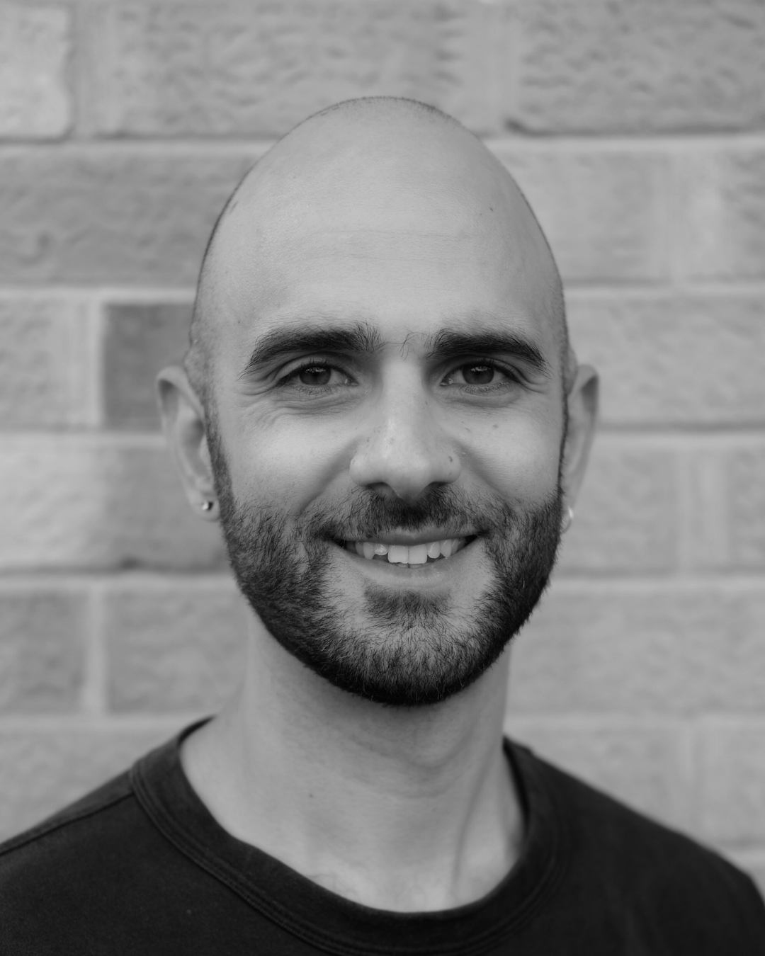 Smiling bald man with a beard in front of a brick wall.