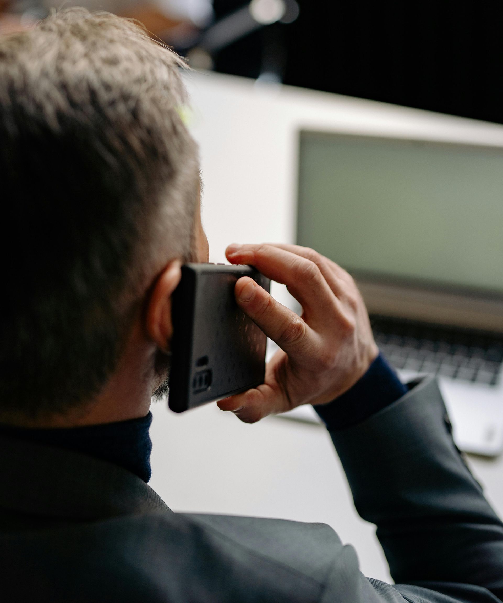 Man in suit on phone, ear to a dark phone. Laptop on a white desk is in background.