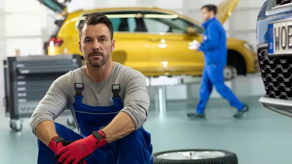 Um mecânico de macacão azul está sentado em uma oficina mecânica com um carro amarelo e outro funcionário ao fundo.