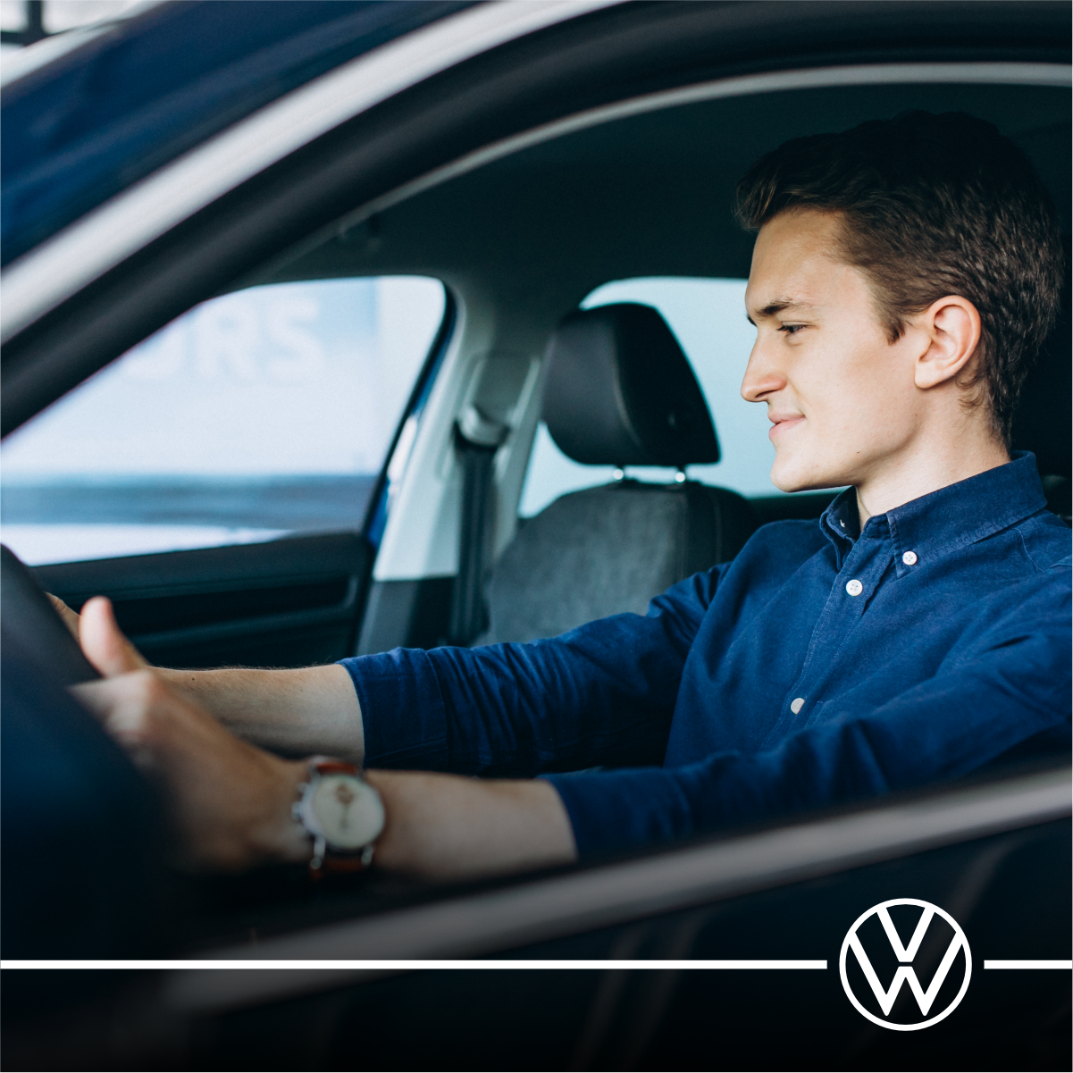 Motorista de camisa azul ao volante de um carro Volkswagen, visto de lado.