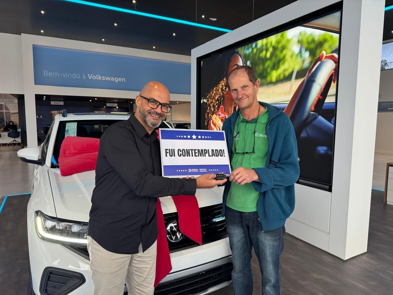Dois homens posando com uma placa em frente a um carro branco em uma concessionária de automóveis.