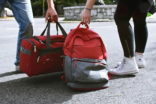 Duas pessoas carregando uma mochila vermelha e uma mochila no asfalto, prontas para viajar.