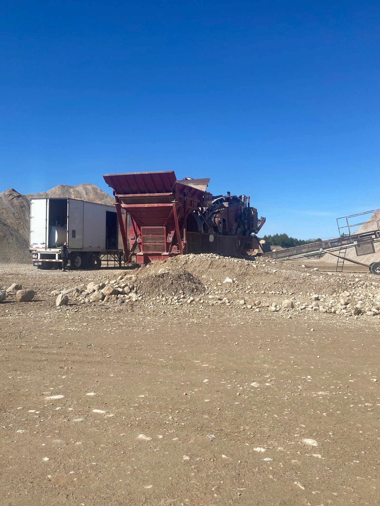 Red machinery with a white trailer in a rocky, outdoor setting under a blue sky.