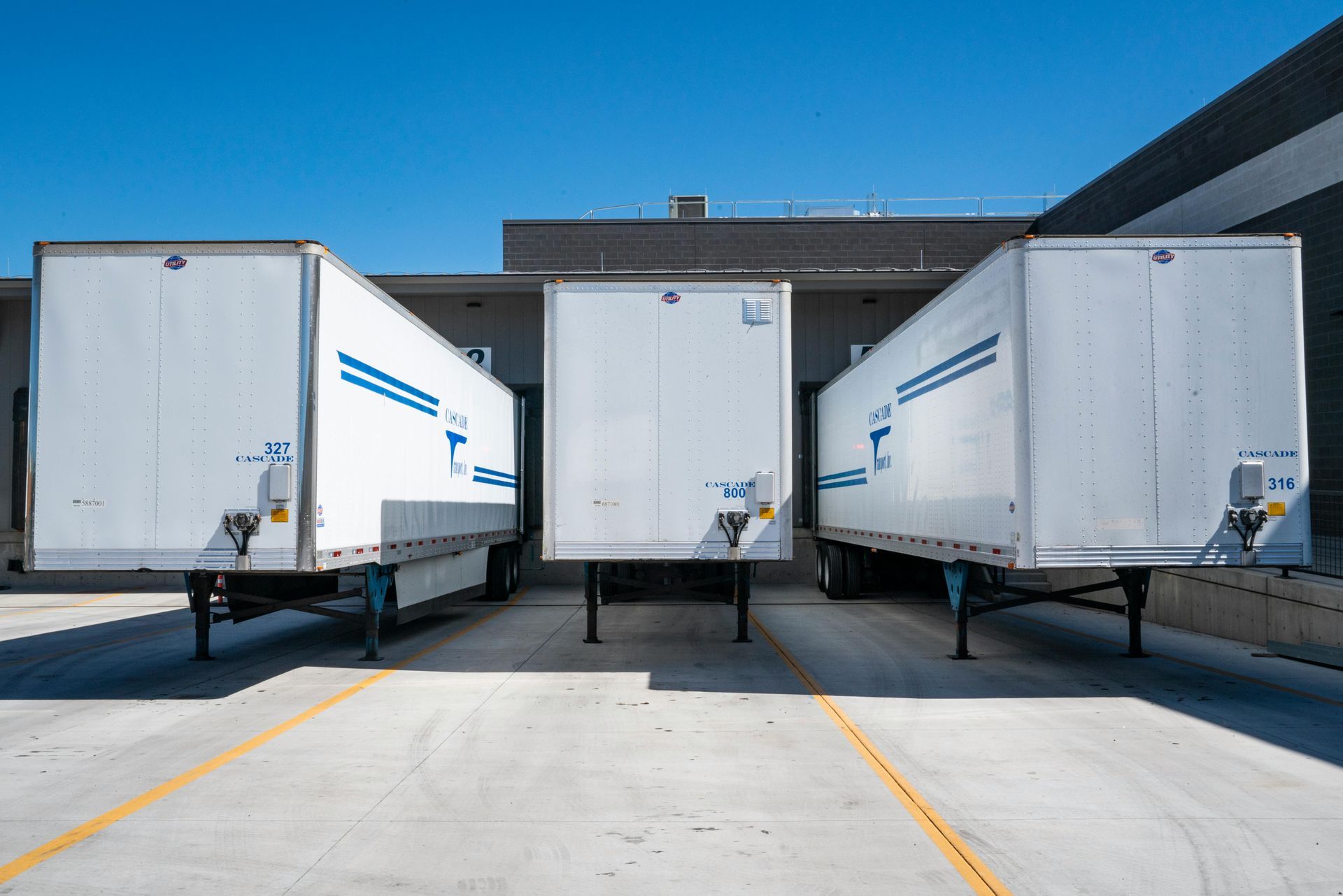 Three white semi-trailer trucks backed up to a loading dock under a blue sky.