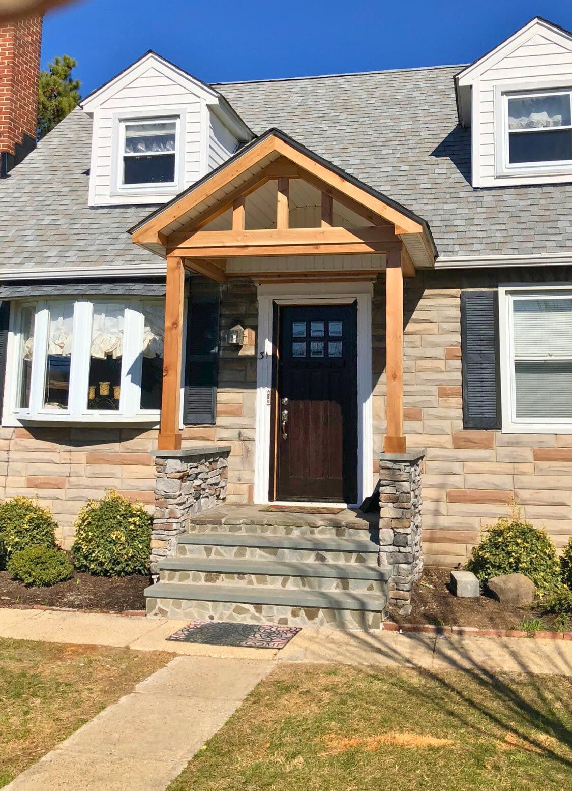 Front entrance of a stone house with a wooden porch, gray roof, and steps leading to the door