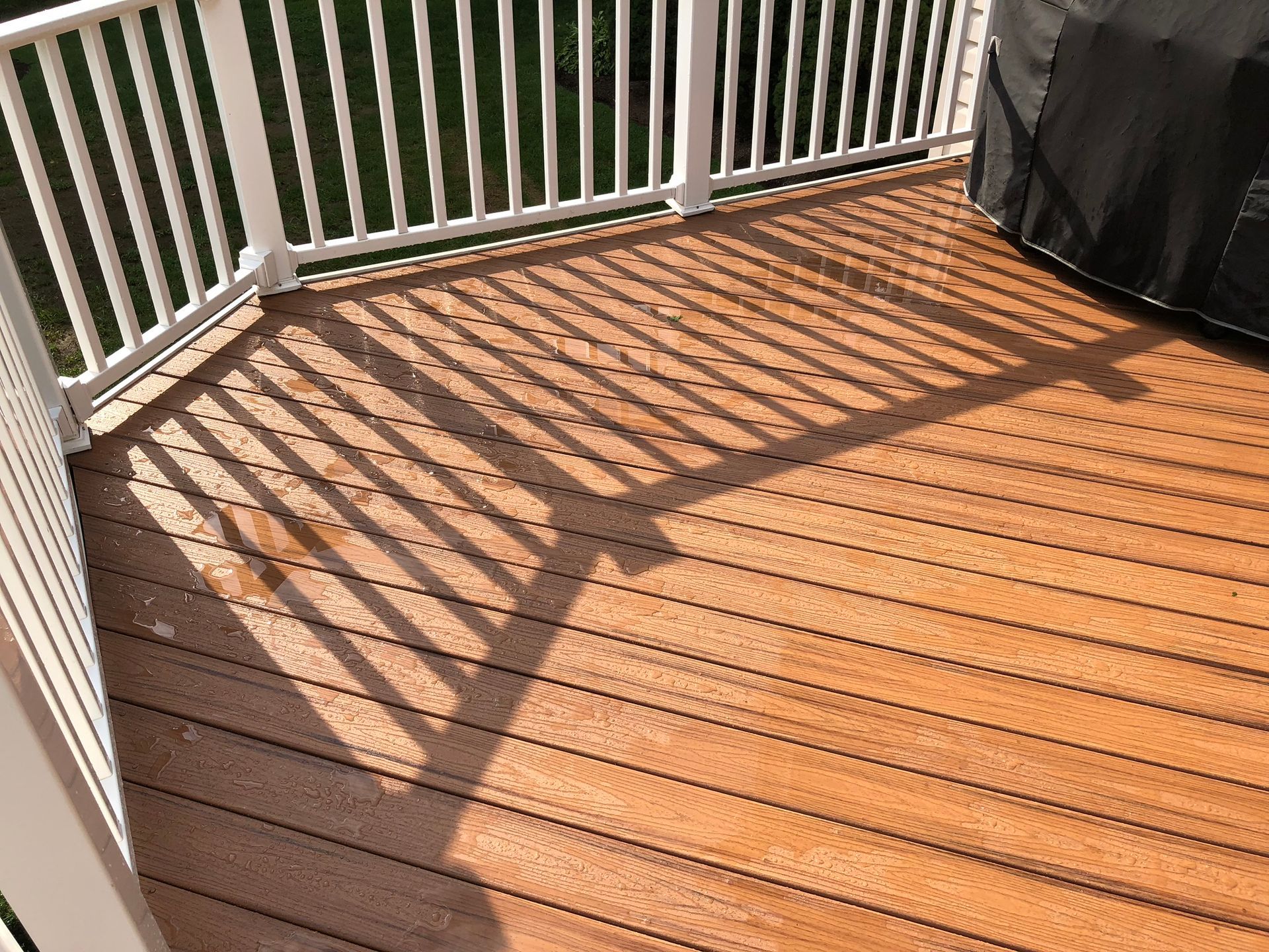 Wooden deck with white railing and diagonal shadows in bright sunlight, beside a covered grill.