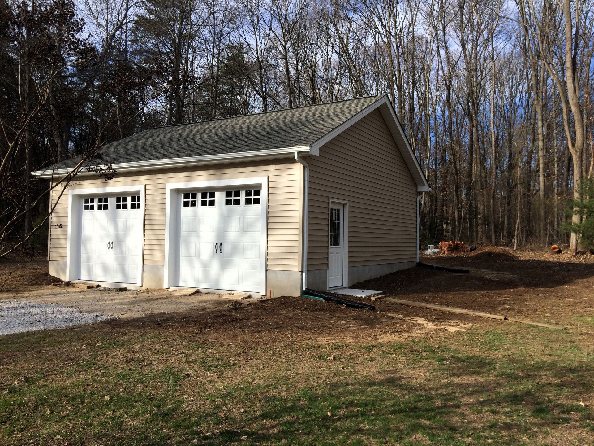 Detached beige two-car garage with white doors beside a wooded yard