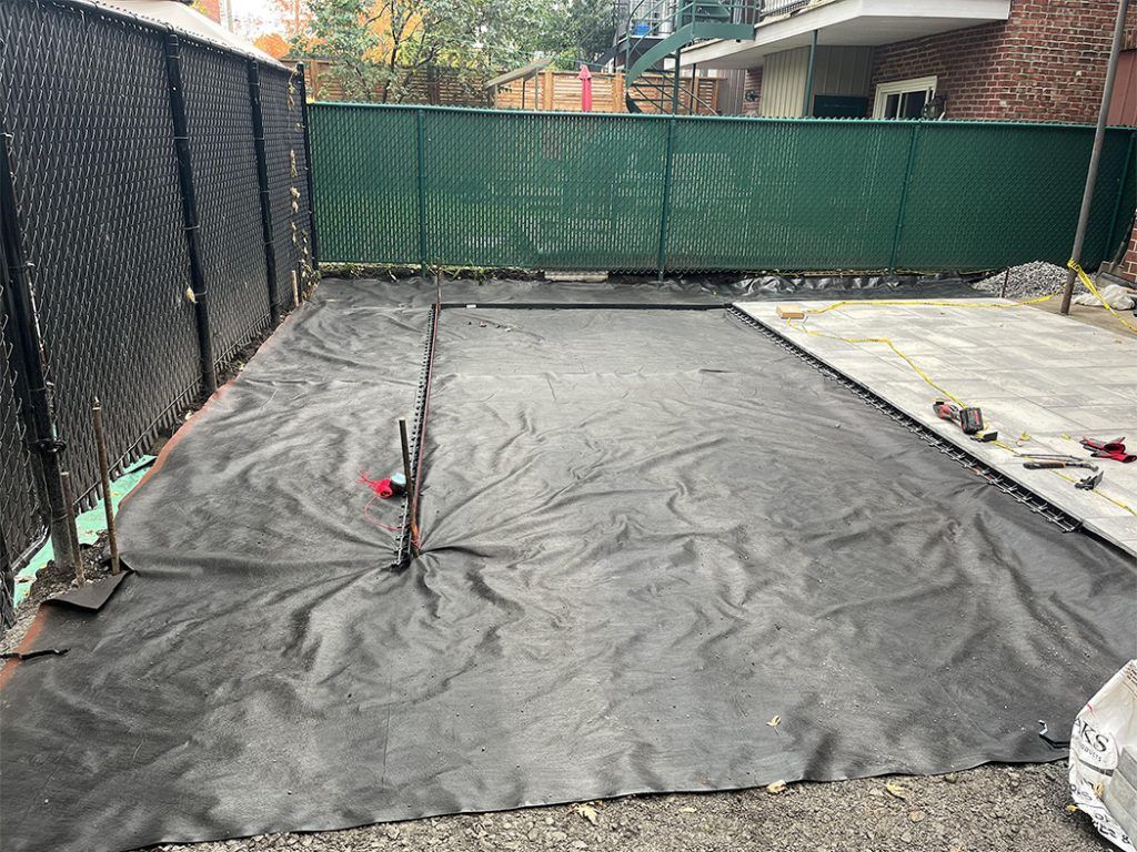 A black tarp is laying on the ground in a backyard next to a chain link fence.
