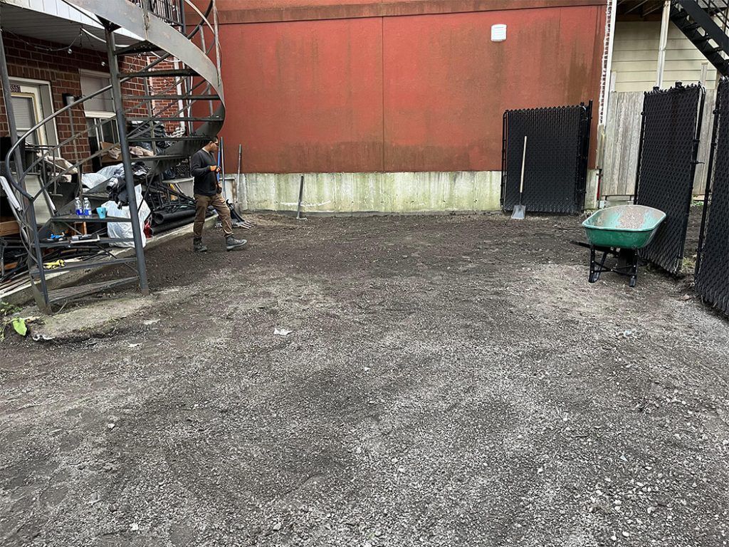 A man is standing in a gravel yard next to a staircase and a wheelbarrow.
