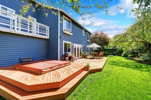 Blue house with wooden deck, hot tub, and patio set beside a green backyard under a bright sky