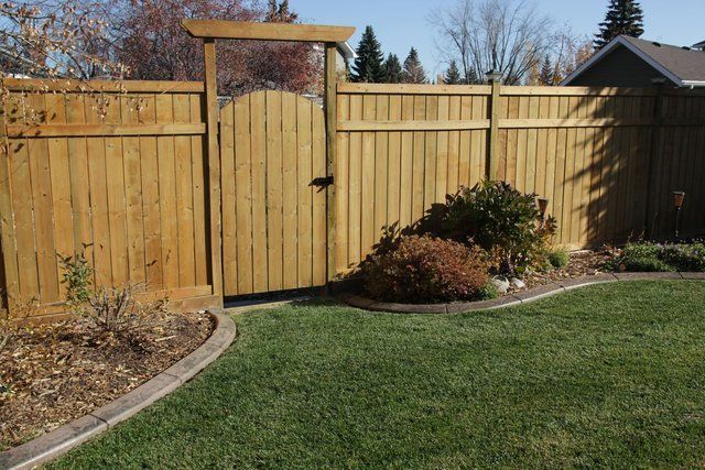 Wooden backyard fence and gate with arched trellis beside a green lawn and flower bed.