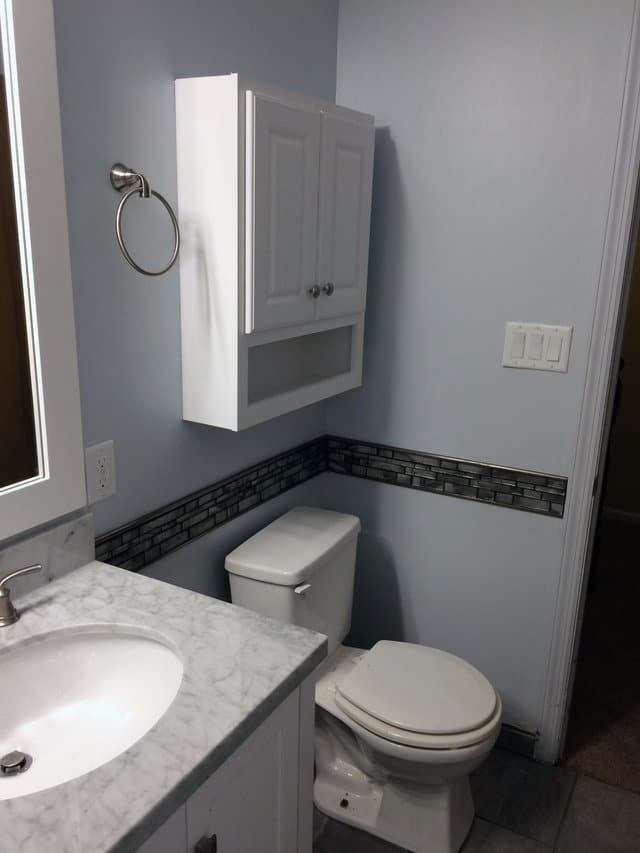 Small bathroom with toilet, sink, mirror, and white wall cabinet over gray tile trim.