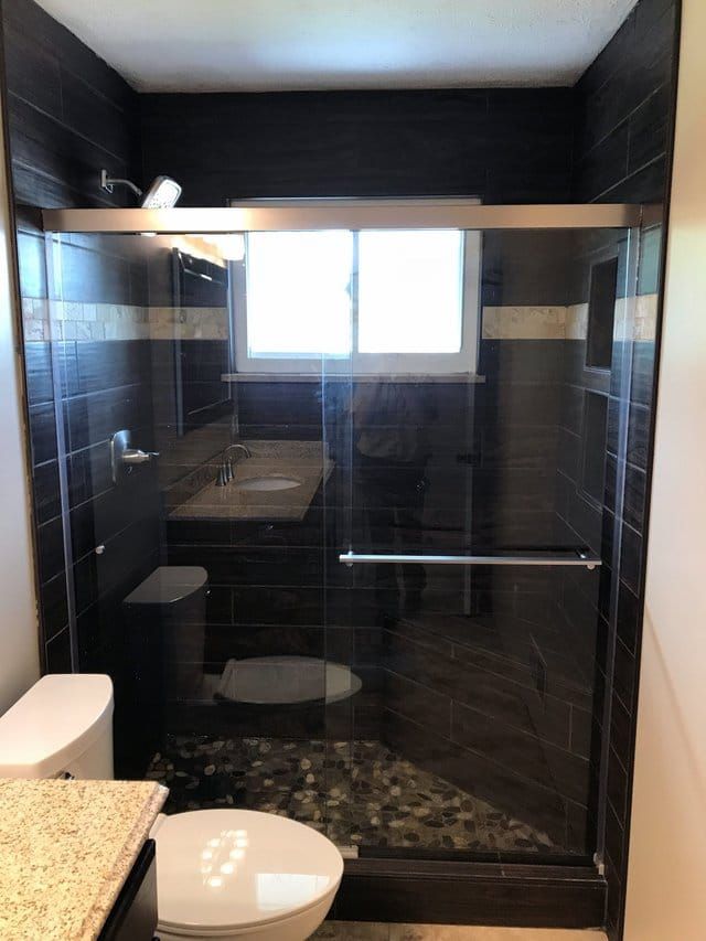 Small bathroom with glass shower enclosure, dark tiled walls, and a window above the sink.