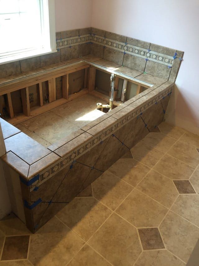 Tiled built-in bathtub under construction in a beige-tiled bathroom near a window.