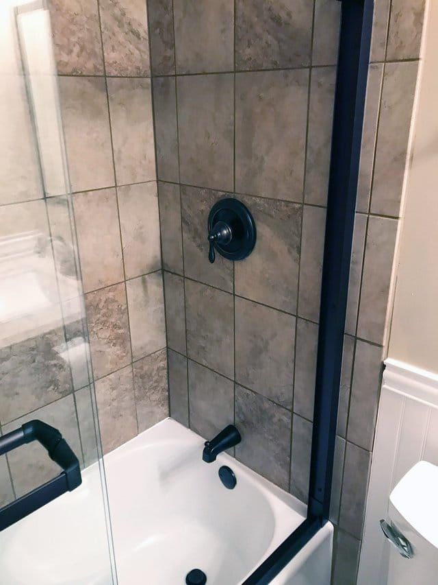 Tiled shower with black fixtures and a glass door, viewed above a white bathtub.