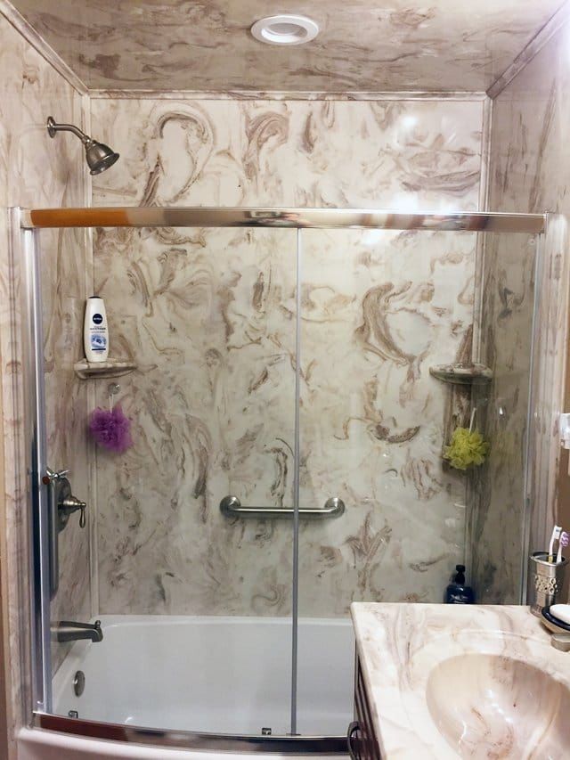 Marble-tiled shower with glass doors, wood shelf, and handheld showerhead in a bathroom