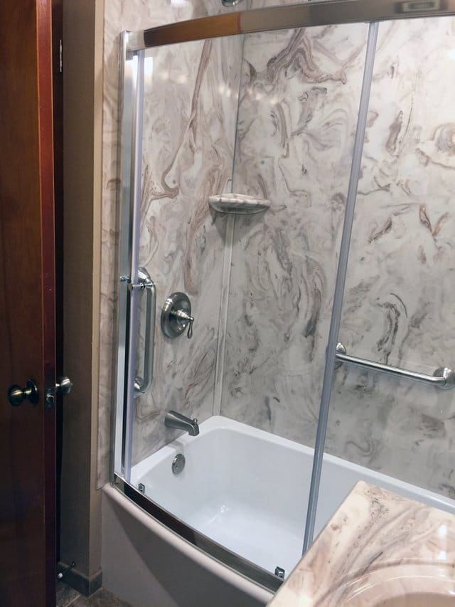 Small bathtub and shower with marble tile walls, glass door, and chrome grab bars in a bathroom