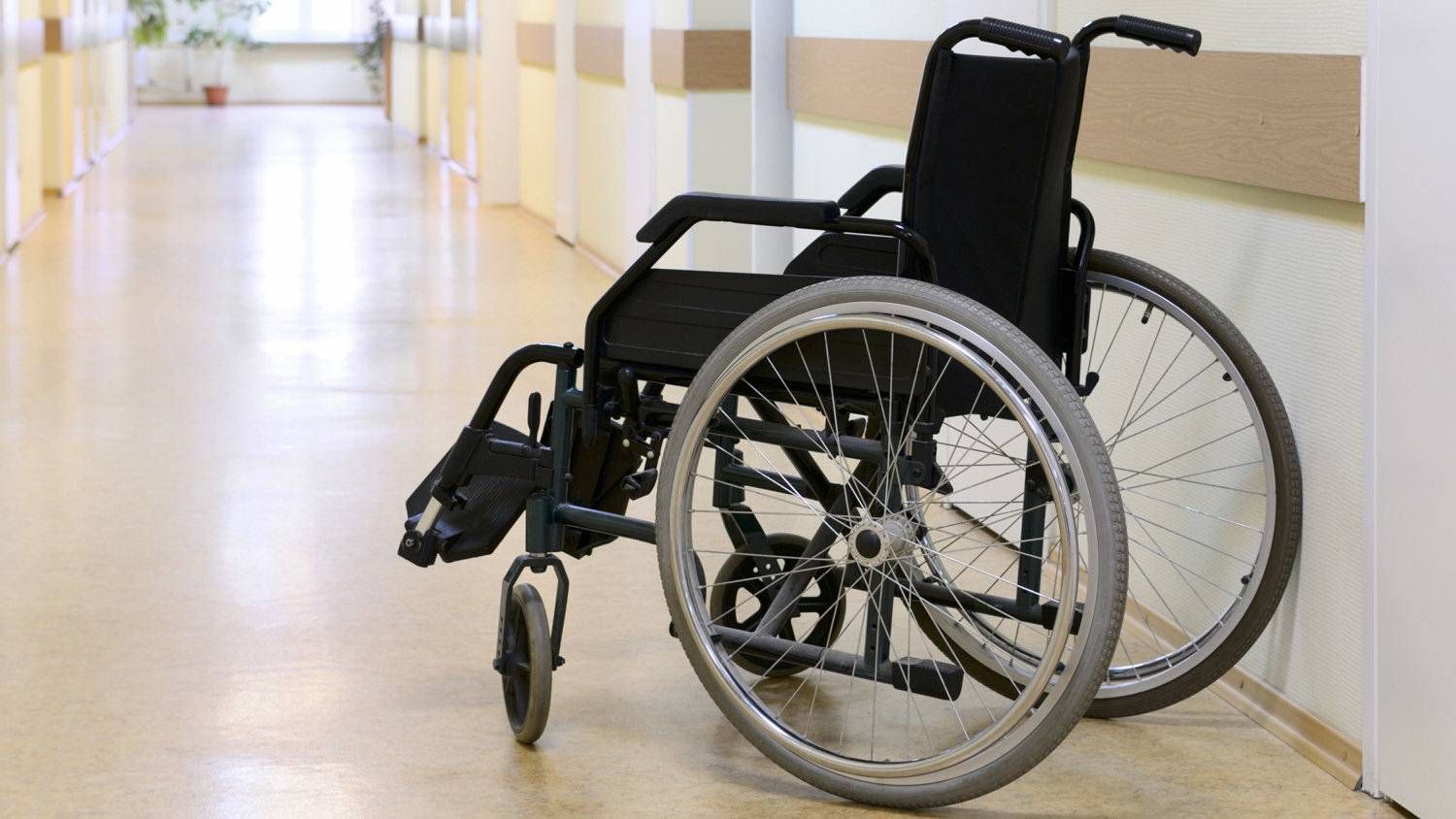 Empty wheelchair parked in a hospital hallway beside a wall.