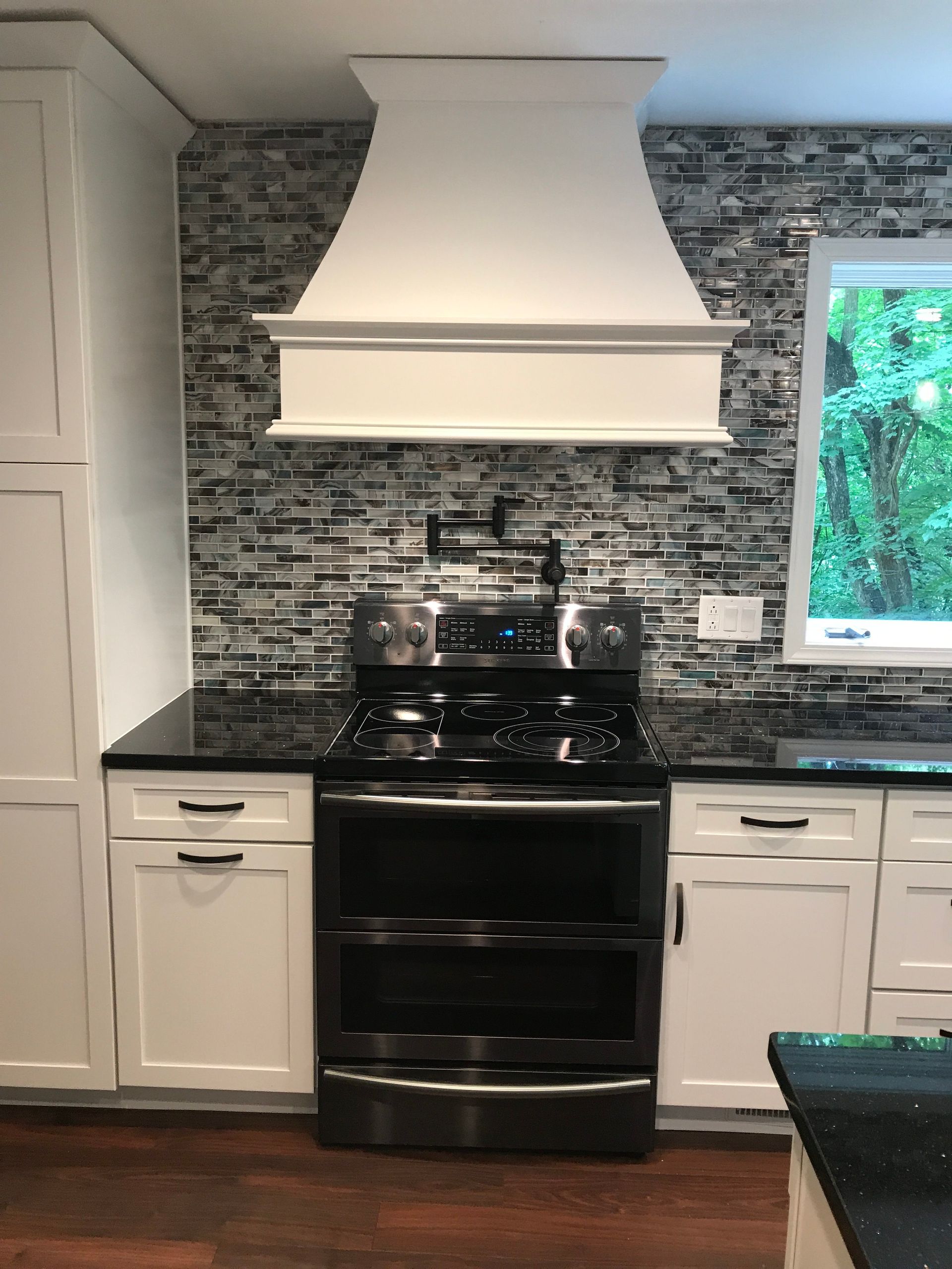 Kitchen with black stove, white cabinets, dark countertops, stone backsplash, and white range hood.