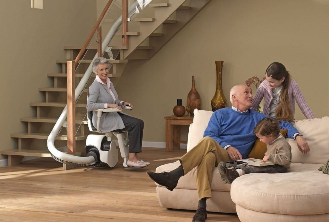People relaxing in a modern living room, with one person seated on a stairlift near the staircase.