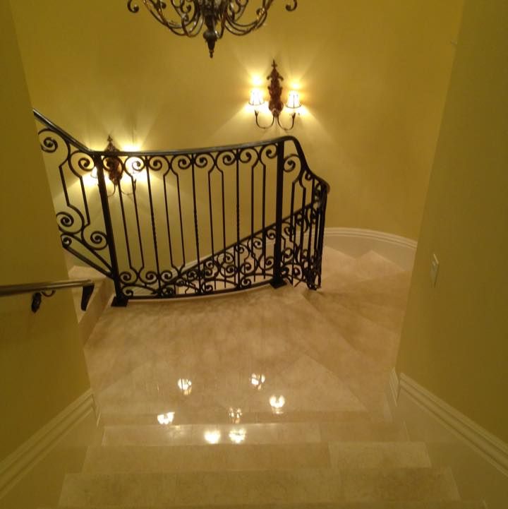 Downward view of a marble staircase with ornate wrought iron railing, lit by sconces and chandelier.