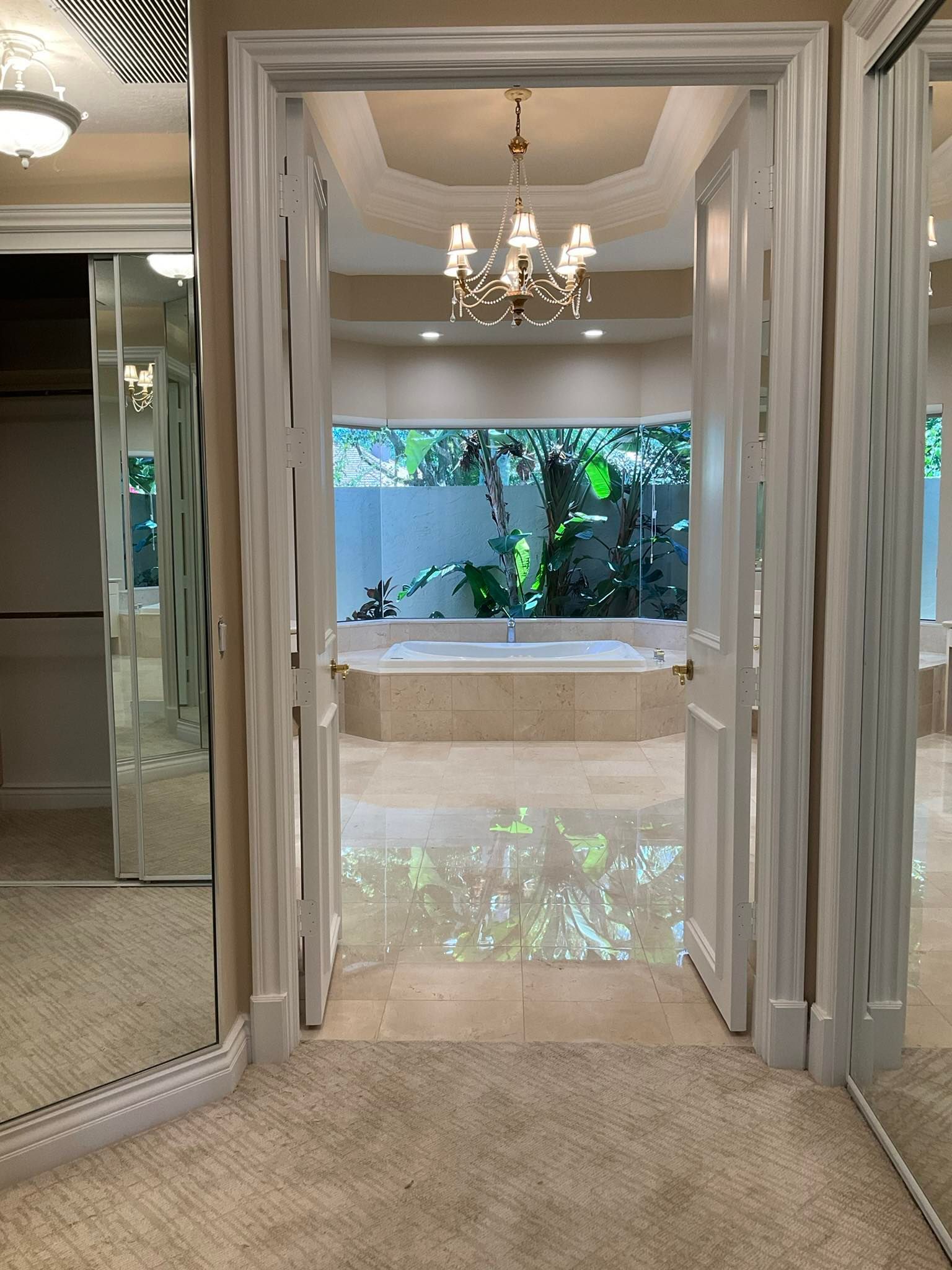 Bathroom doorway with view of a tub and window. Light beige tile and walls, chandelier, and a mirror wall.