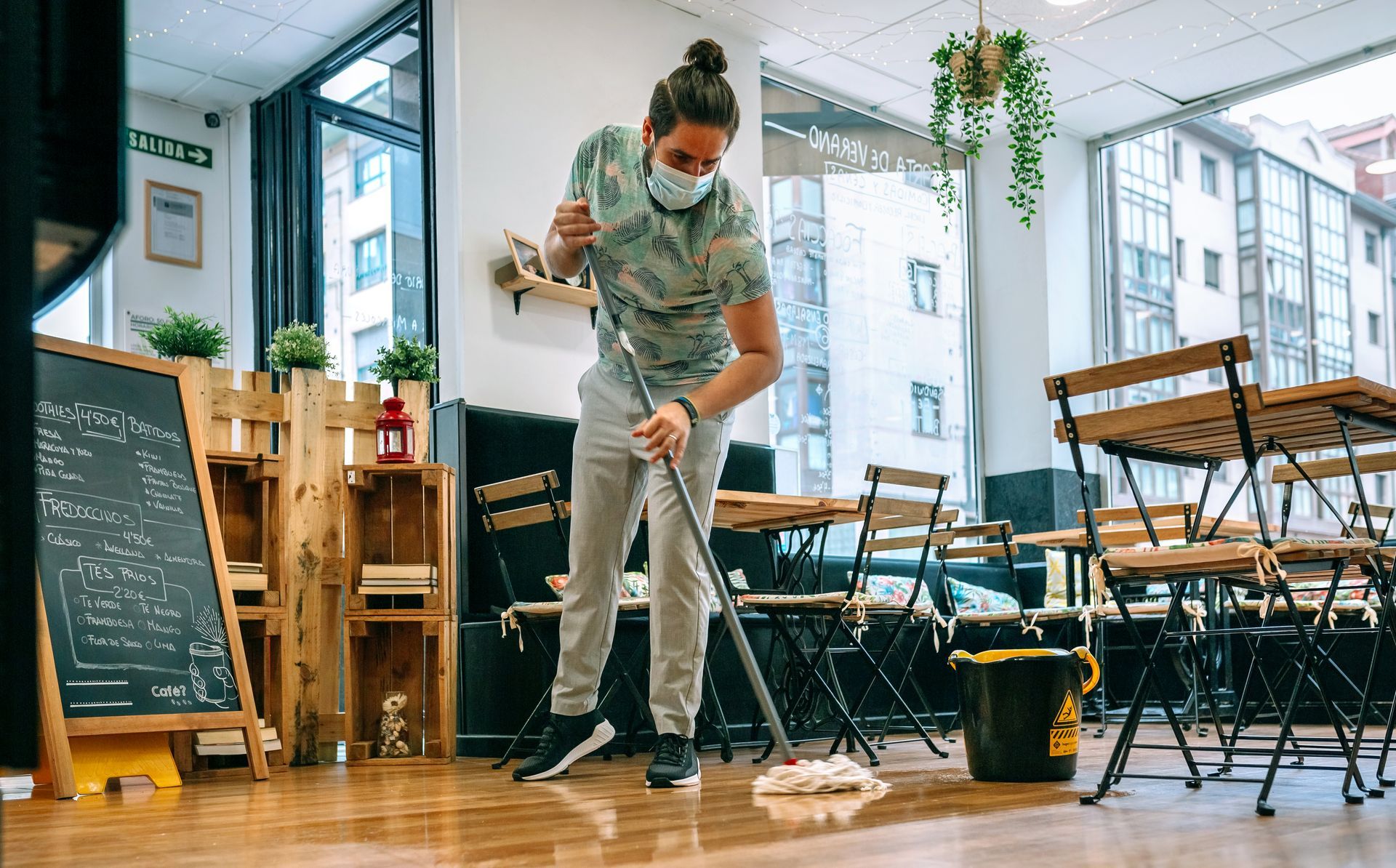 Person wearing a mask mopping the floor of a cafe; tables and chairs visible.