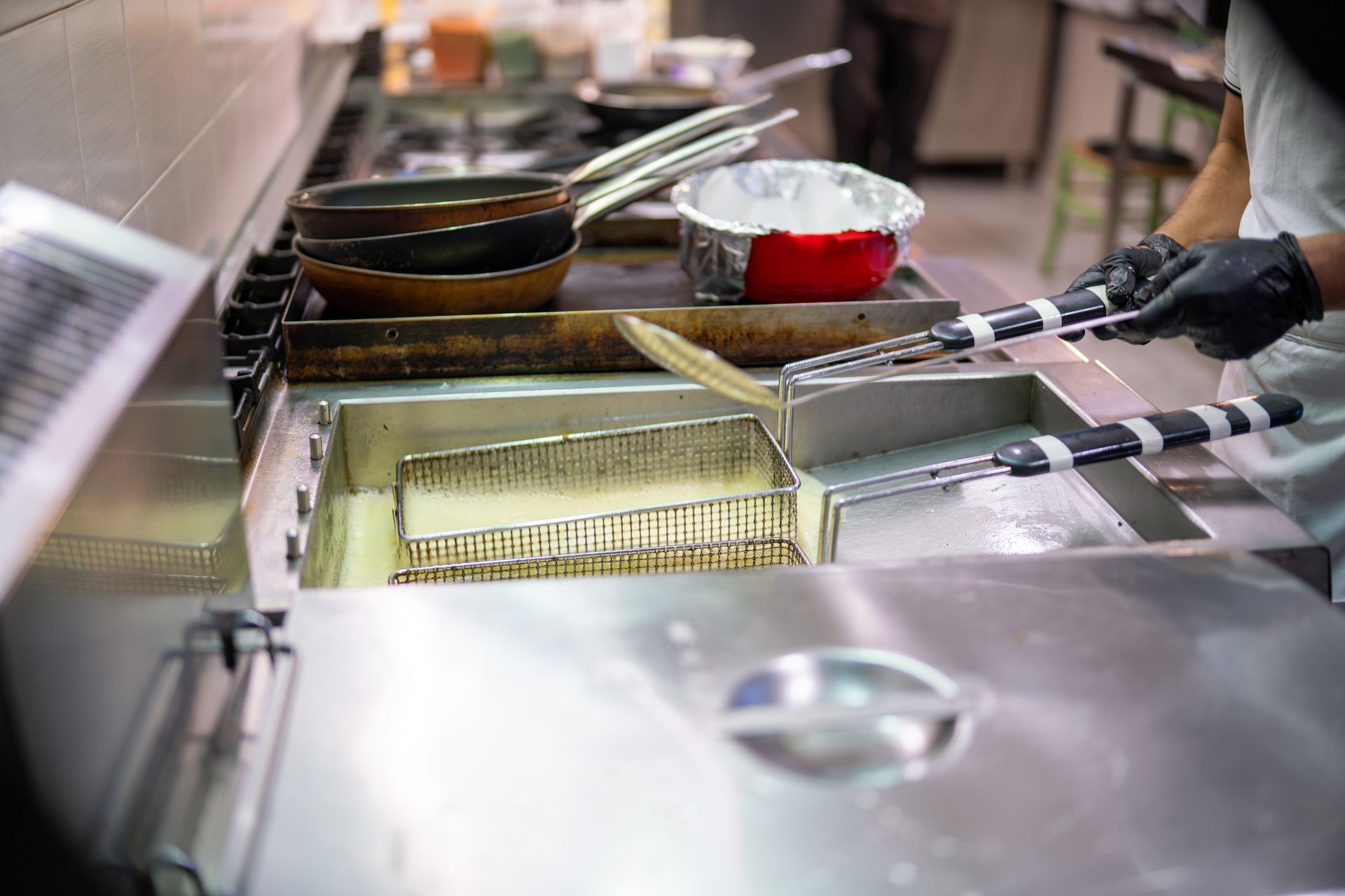 Chef using a deep fryer in a commercial kitchen; black gloved hands, metal, and cooking equipment.