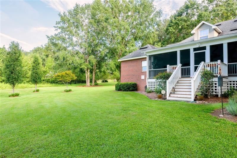 A red brick house with a screened-in porch and white wooden stairs, overlooking a large, lush green lawn with trees.