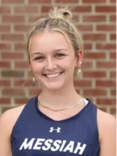 A girl wearing a messiah jersey is smiling in front of a brick wall.