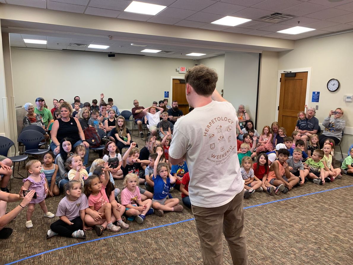 Presenter facing a seated crowd in a large room with many children and adults listening.