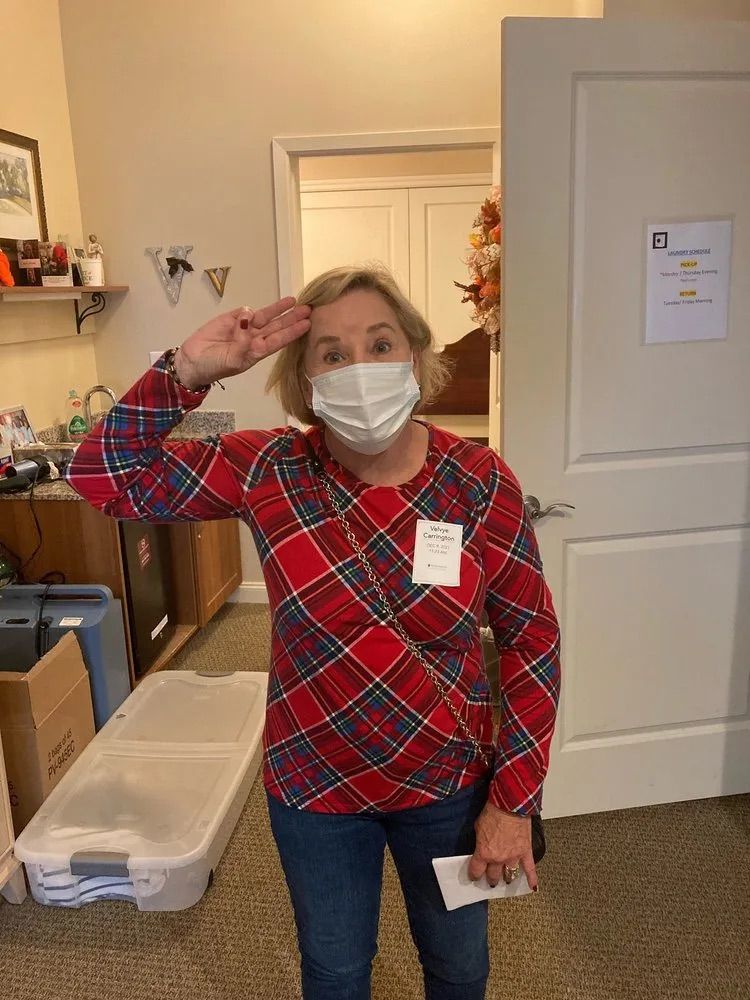 A person in a red plaid shirt and white face mask salutes while standing in an office with an open door.
