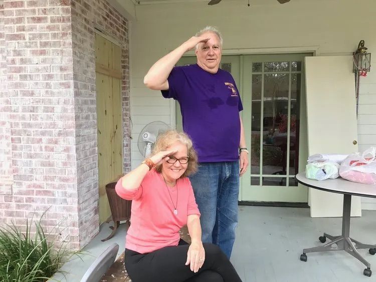 Two people stand on a porch, both smiling and saluting with their right hands raised to their foreheads.