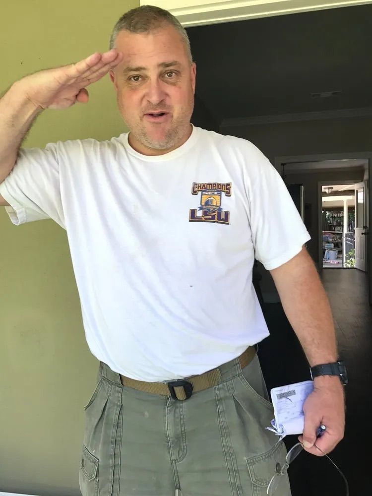 A man in a white t-shirt and olive green shorts salutes while holding a small object in his left hand.