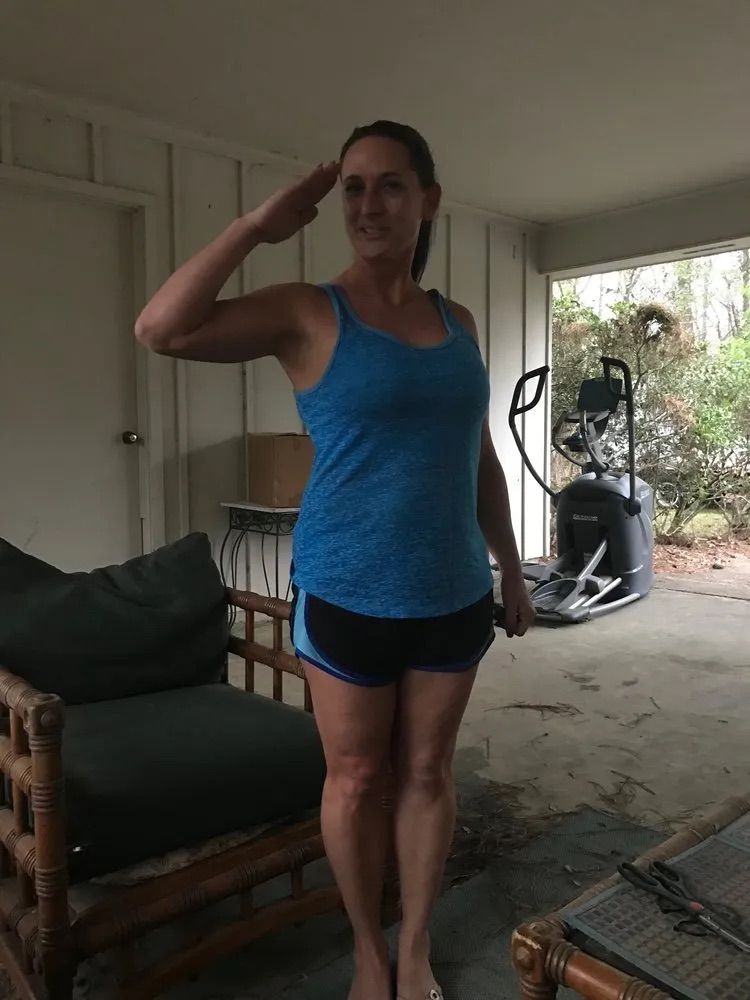 A person wearing a blue tank top and dark athletic shorts stands on a patio and gives a salute.