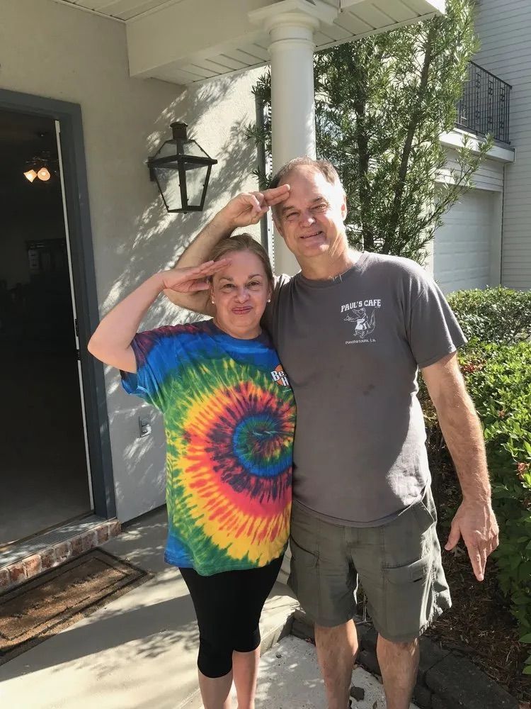Two people stand on a porch and offer a salute toward the camera.