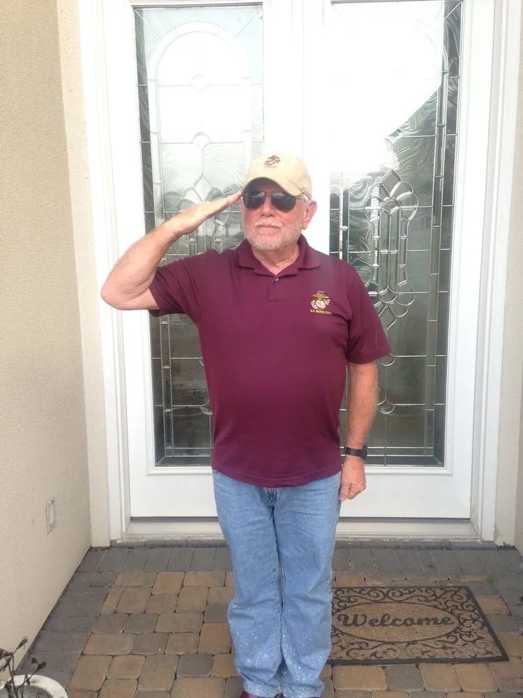 A person wearing a maroon polo shirt, blue jeans, and a baseball cap standing outside and giving a salute.
