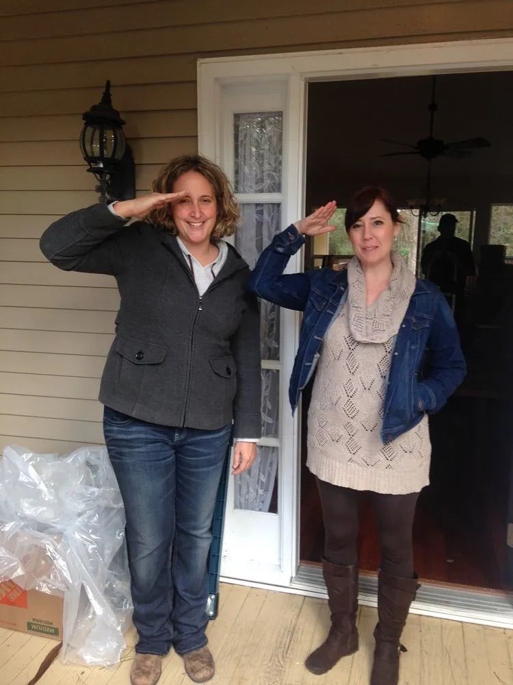 Two people standing on a porch, both smiling and giving a salute gesture, wearing casual jackets and sweaters.